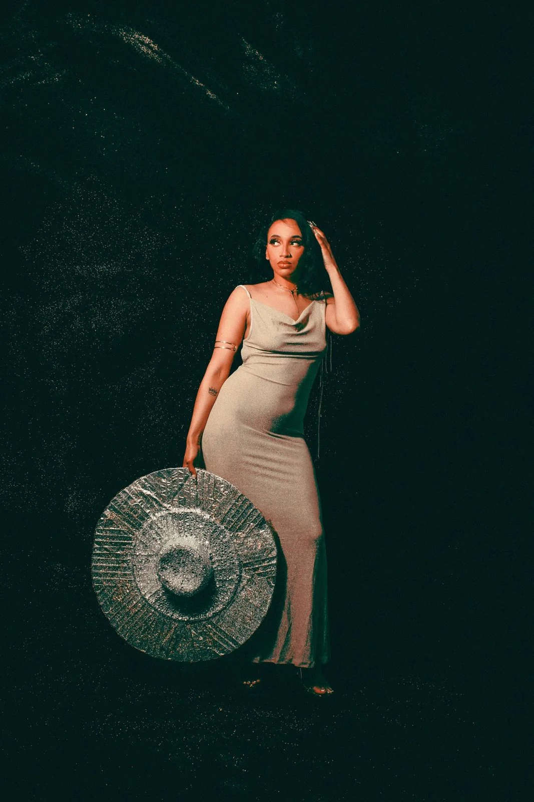 A woman in a light dress stands before a dark sparkling backdrop holding a circular silver textured piece